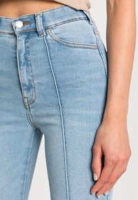 Ljusblå högmidjade jeans med en slät textur, kontrastsömmar och en smal passform. Har en framknäppning och femficksdesign.