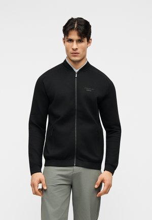 Muž v čiernej bomberyškej bunde na zips a sivých nohaviciach, stojaci pred jednoliatym svetlým pozadím, čelom k fotoaparátu s neutrálnym výrazom.