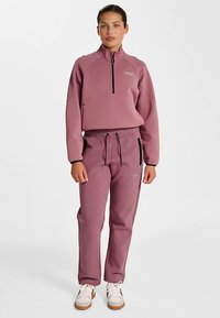 Paarse fleece trui met halve ritssluiting, geribde hals en bijpassende joggers met trekkoorden en zijzakken, gecombineerd met witte sneakers.