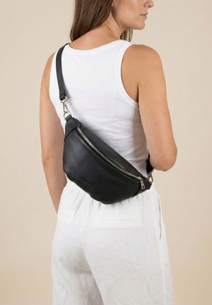 Vrouw draagt een witte mouwloze top en broek met een zwarte leren crossbodytas over haar rug.