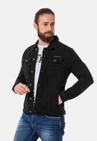Veste en denim noir avec un col classique, fermeture à boutons, deux poches poitrine et un léger détail de marque sur la poitrine. Portée sur une chemise blanche.
