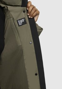 Olijfgroene, gevoerde jas met zwarte accenten aan de binnenkant. Bevat een merketiket en drukknoopsluiting. Glad textuur, modern ontwerp.