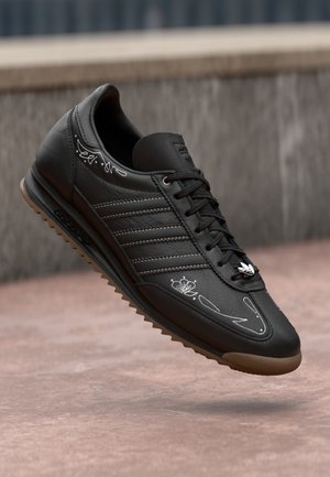 Zwarte leren sneaker met bruine zool, witte bloemstiksels, drie zijstrepen en metalen logo op de veters, getoond zwevend boven de grond.