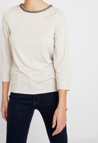 Haut à manches longues crème avec un accent texturé au niveau du col. Tissu doux, ourlet rond et manches mi-longues. Associé à un jean en denim sombre.