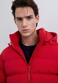 Joven con cabello oscuro que lleva una chaqueta acolchada roja con cremallera sobre un suéter negro, mirando directamente a la cámara frente a un fondo sencillo.