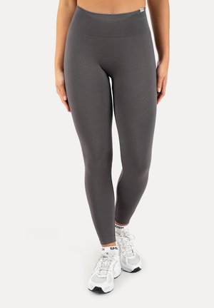 Femme portant des leggings sans coutures gris foncé taille haute et des chaussures de sport blanches, debout, les mains détendues le long du corps.