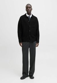 Melna adīšanas kardigāna ar lielām pogām un krūšu kabatu, apvienots ar pelēkiem biksēm un melnām kurpēm uz neitrāla fona.