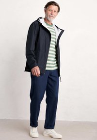 Lättviktig svart jacka med vit huva, kombinerad med en grönrandig t-shirt och marinblå byxor. Vita sneakers fulländar outfiten.