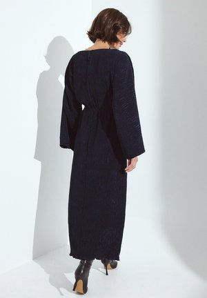 Langes, marineblaues Maxikleid mit strukturiertem, plissiertem Design, weiten Ärmeln und einem Kordelzug in der Taille. Getragen mit schwarzen High-Heels.