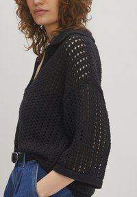 Zwarte gebreide trui met een losse, open ontwerp met een V-hals, verlaagde schouders en ribbelboorden. Draagt over blauwe jeans.