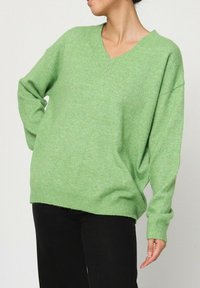 Pull vert en V, fabriqué en maille douce, avec des manches amples et une silhouette décontractée. Tissu texturé avec une légère variation.