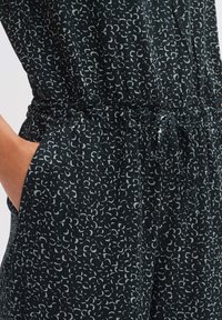 Schwarzer Jumpsuit mit wiederholtem, hellgrauem abstraktem Muster. Verfügt über einen elastischen Bund und Taschen. Weicher Stoff und lockere Passform.