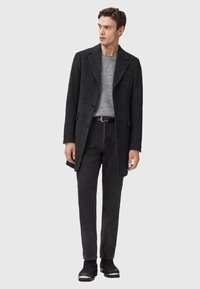 Svart, strukturerad kappa med krage, bärs över en grå tröja, kombinerat med svarta jeans och ankelstövlar. Klassisk, skräddarsydd silhuett.