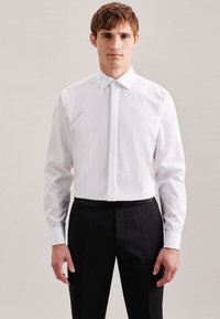 Uomo che indossa una camicia bianca aderente con bottoni nascosti e pantaloni eleganti neri su uno sfondo semplice.