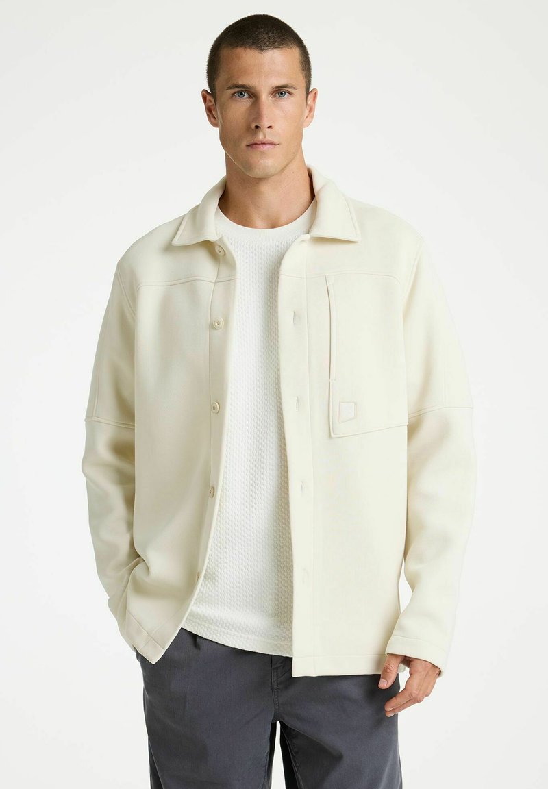 Crème-kleurige jas met een relaxed fit, voorknopen en een enkele borstzak, gedragen over een gestructureerd wit shirt en grijze broek.