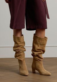 Suède enkellaarsjes met een slouchy design, tan kleur, puntige neus en blokhak. Draag je met bordeaux cropped broeken, op een houten vloer.