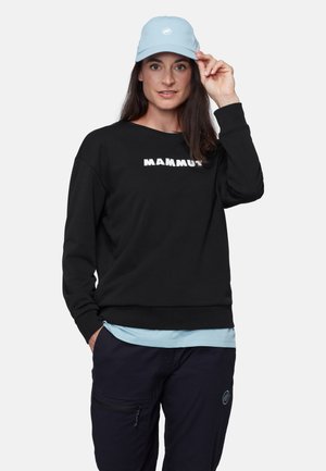 Kobieta w czarnej bluzie Mammut, jasnobłękitnej czapce i ciemnych spodniach, pozująca z jedną ręką regulującą daszek czapki.