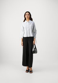 Camicia bianca con colletto e tasca sul petto, maniche arricciate, abbinata a pantaloni neri a pieghe a gamba larga e una borsa nera a tracolla.