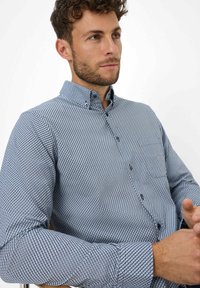 Camicia a bottoni blu e bianca con un motivo, colletto, tessuto texturizzato, maniche lunghe e bottoni scuri, indossata da una figura maschile seduta.