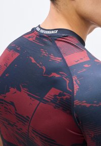 Κοντινό πλάνο του πάνω μέρους της πλάτης και του λαιμού ενός ατόμου που φοράει ένα σφιχτό μπλουζάκι με μοτίβο σε μπλε ναυτικό και κόκκινο, με τη λέξη "PERFORMANCE" στο γιακά.