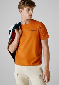Orange bomulds t-shirt med rund halsudskæring og korte ærmer. Har "RED BULL RACING" trykt i sort. Kombineret med lyse farvede cargo bukser.