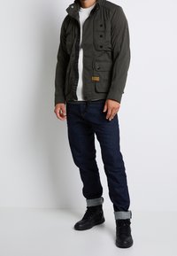 Zielona oliwkowa kurtka robocza z wieloma kieszeniami, czarnymi guzikami i wysokim kołnierzem, noszona na białym swetrze z prążkowanej dzianiny oraz ciemnych dżinsach. Czarny obuwie.