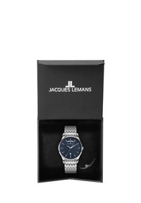 Reloj plateado con una correa de acero inoxidable estampada, esfera azul, marcadores de hora plateados y pantalla de fecha, alojado en una caja negra con el logotipo.