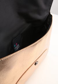 Kultainen metallinhohtoinen käsilaukku, jossa on sileä pinta, musta kangasvuori ja läppäsuljin. Sisältää näkyvän merkkitagin sisällä.