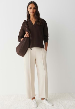 Pullover marrone con scollo a V e dettaglio di arricciatura sulle spalle, abbinato a pantaloni crema a gamba larga e sneaker bianche. Stile minimalista.