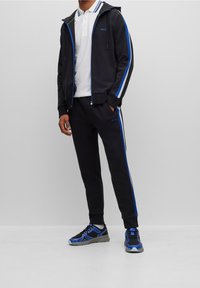 Survêtement noir avec des rayures latérales bleues et blanches, comprenant un sweat à capuche zippé et un pantalon à taille élastique. Associé à des baskets sportives bleues.