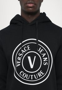 Must mustaga, mis on valmistatud puuvillast, millel on suur valge ümmargune logo, millel on tekst "VERSACE JEANS COUTURE" ja silmatorkav "V" keskel.