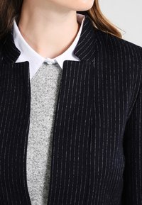 Laivastonsiniset liituraitableiseri, jossa on räätälöity kaulus, kerrospukeutuneena vaaleanharmaan neuleen päälle. Ominaisuuksiin kuuluu sileä pinta ja siistit linjat.