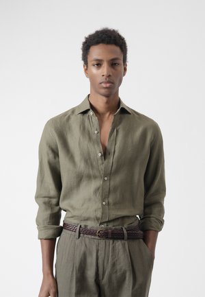 Joven con camisa de lino verde oliva con las mangas remangadas y pantalones a juego, cinturón trenzado oscuro, de pie frente a un fondo liso.