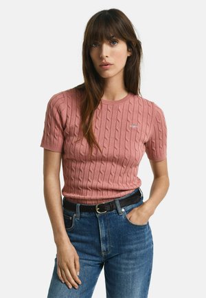 Cable-knit short-sleeve sweater in soft pink, featuring a round neckline and subtle logo detail, paired with high-waisted blue jeans and a black belt.