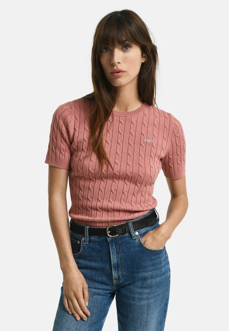 Kabelgebreide trui met korte mouwen in zachtroze, met een ronde halslijn en subtiele logodetails, gecombineerd met high-waisted blauwe jeans en een zwarte riem.