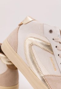 Vita och beige sneakers med metalliska guldaccenter; har texturerat läder och mocka, rundad tå och platt gummisula.