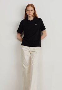 Schwarzes Baumwoll-T-Shirt mit kurzen Ärmeln und lockerer Passform, mit einem kleinen grünen Logo auf der Brust, kombiniert mit off-white weit geschnittenen Hosen.