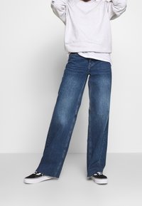 Modré džínové kalhoty s širokými nohavicemi, s vybledlým efektem, klasickým pětikapsovým střihem a jemným šitím podél švů, kombinované s černými teniskami.