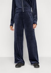 Hose aus samtigem, marineblauem Material mit entspannter Passform, breiten Beinen und glatter Textur, kombiniert mit schwarzen Sportschuhen.