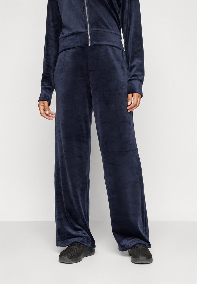 Pantalones de chándal de terciopelo azul marino con un corte relajado, que presentan piernas anchas y una textura suave, combinados con zapatillas deportivas negras.