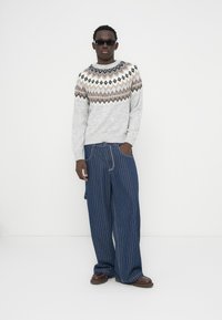 Γκρι πουλόβερ με διακοσμητική λωρίδα σε γήινους τόνους, συνδυασμένο με παντελόνι φαρδύ, σε μπλε σκούρο με ρίγες και σκούρα παπούτσια. Άνδρας που φορά γυαλιά ηλίου.