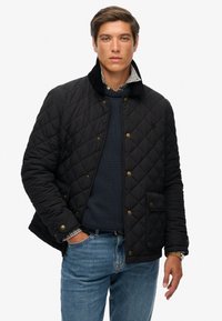 Czarna pikowana kurtka z kołnierzem z materiału sztruksowego, zatrzaskami i kieszenią z przodu, noszona na niebieskim swetrze i koszuli w kratę. Dżinsowe spodnie.