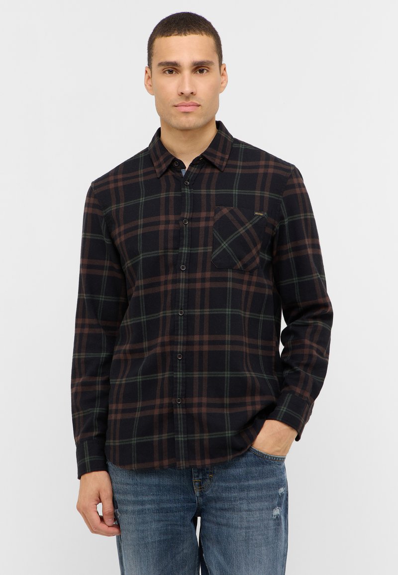 Chemise en flanelle noire avec un motif à carreaux verts et marron, manches longues, col à boutonnière et poche poitrine. Portée avec un jean bleu.