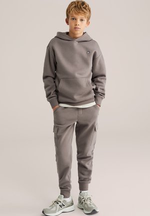 Siva dukserica s kapuljačom i prednjim džepom te izvezenim logotipom, uparen s odgovarajućim cargo hlačama sa bočnim džepovima i rebrastim manžetama, nošena sa sivim tenisicama.