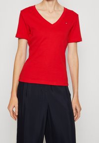 Röd bomulls-T-shirt med V-ringning och korta ärmar, med en liten logotyp på bröstet. Kombinerad med löst sittande marinblå byxor.