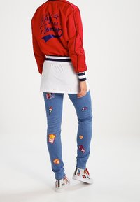 Czerwona kurtka bombka z logo, biała koszulka nałożona pod spód, niebieskie dżinsy z kolorowymi łatami i białe sneakersy z akcentami.