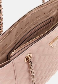 Light pink quilted handbag with gold hardware, chain strap, and patterned interior featuring repeating 'GUESS' logo. Zippered compartments visible.