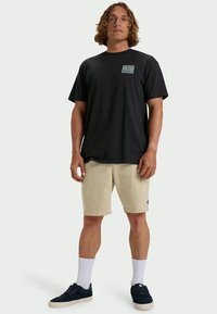 Man with shoulder-length curly hair wearing a black T-shirt with a small turquoise wave logo, beige shorts, white socks, and black sneakers.