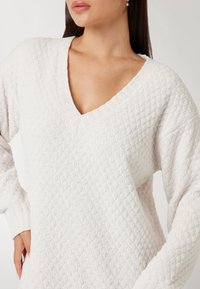 Witte gebreide trui met een V-hals, een textuurpatroon en losse mouwen, gemaakt van een zacht en dik materiaal voor extra warmte.