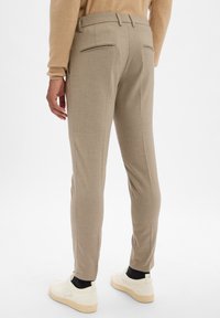 Pantalons beige sur mesure avec deux poches zippées à l'arrière, coupe slim, et une texture lisse, portés avec des baskets blanches et des chaussettes noires.